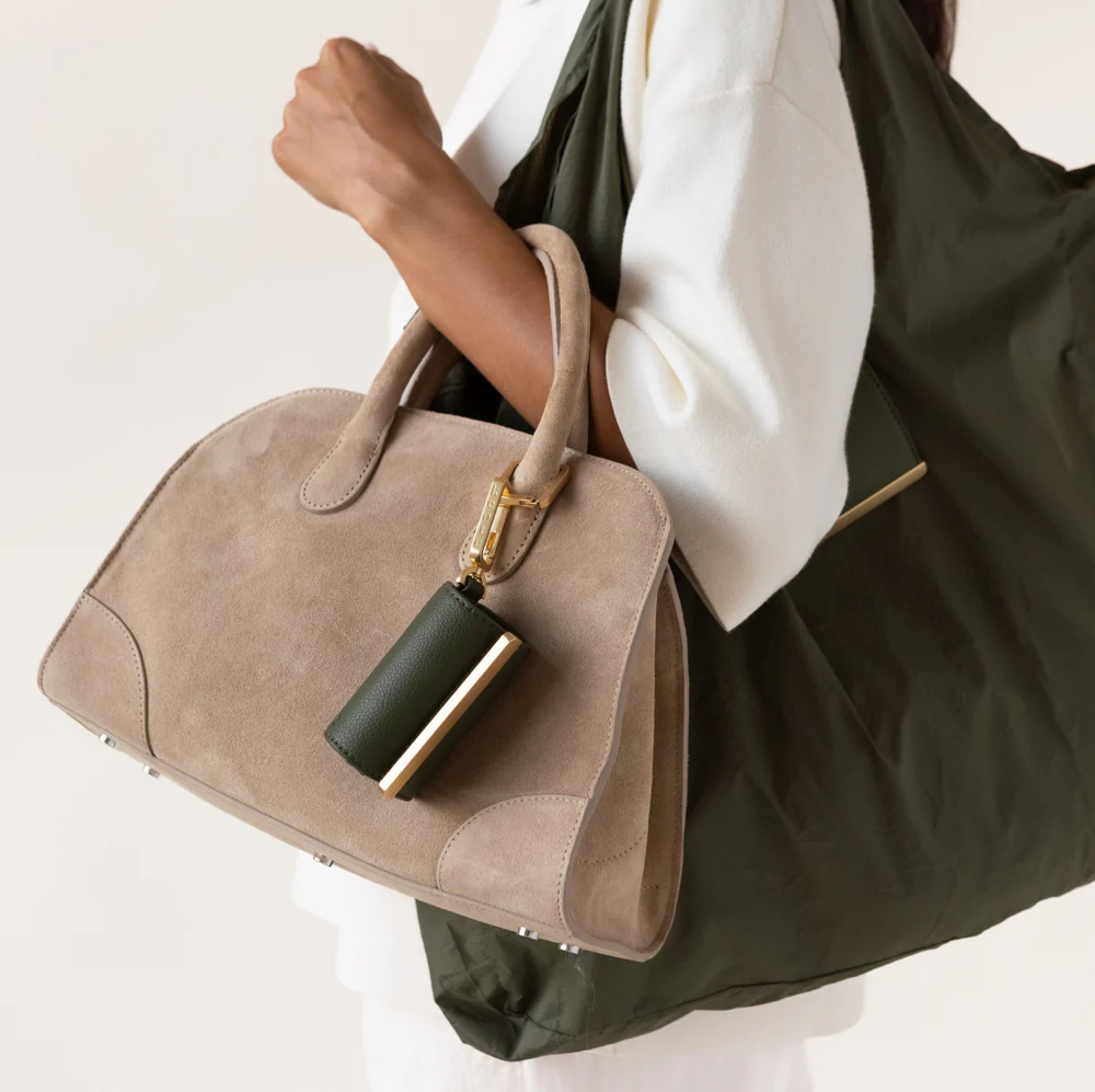A person carries a soft olive-green foldable tote bag over one shoulder while holding a structured taupe suede handbag. Attached to the handbag is a matching olive-green leather accessory with gold-tone hardware. The neutral color palette and clean styling create a s