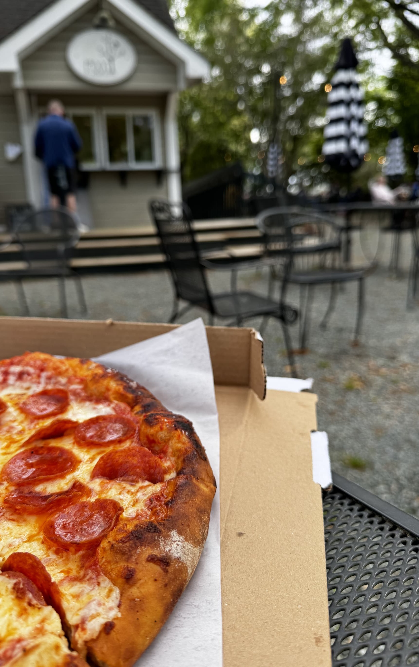 Close-up of a pepperoni pizza in an open box on a metal patio table, with a blurred outdoor café setting in the background. Behind it, a small food stand and scattered tables with black-and-white striped umbrellas sit among trees, suggesting a casual, relaxed dining area.