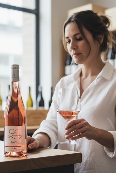 Woman in a light blouse stands at a wine shop counter, holding a glass of rosé while looking at the bottle beside her. Shelves of wine line the softly lit background, creating a calm, refined tasting-room atmosphere.