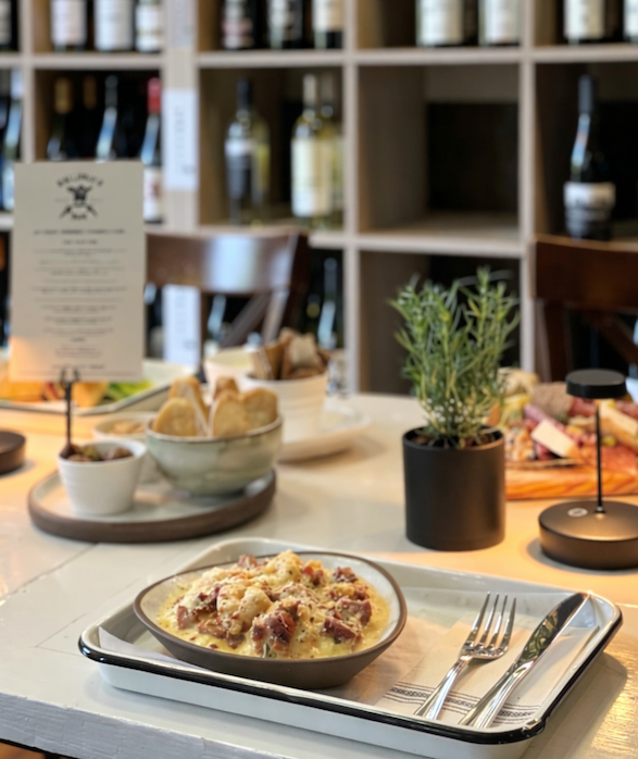 Table set at Galloway's Wine & Beer Bar with a plated pasta dish, bread, herbs, and menus in front of shelves filled with wine bottles, softly lit and inviting.