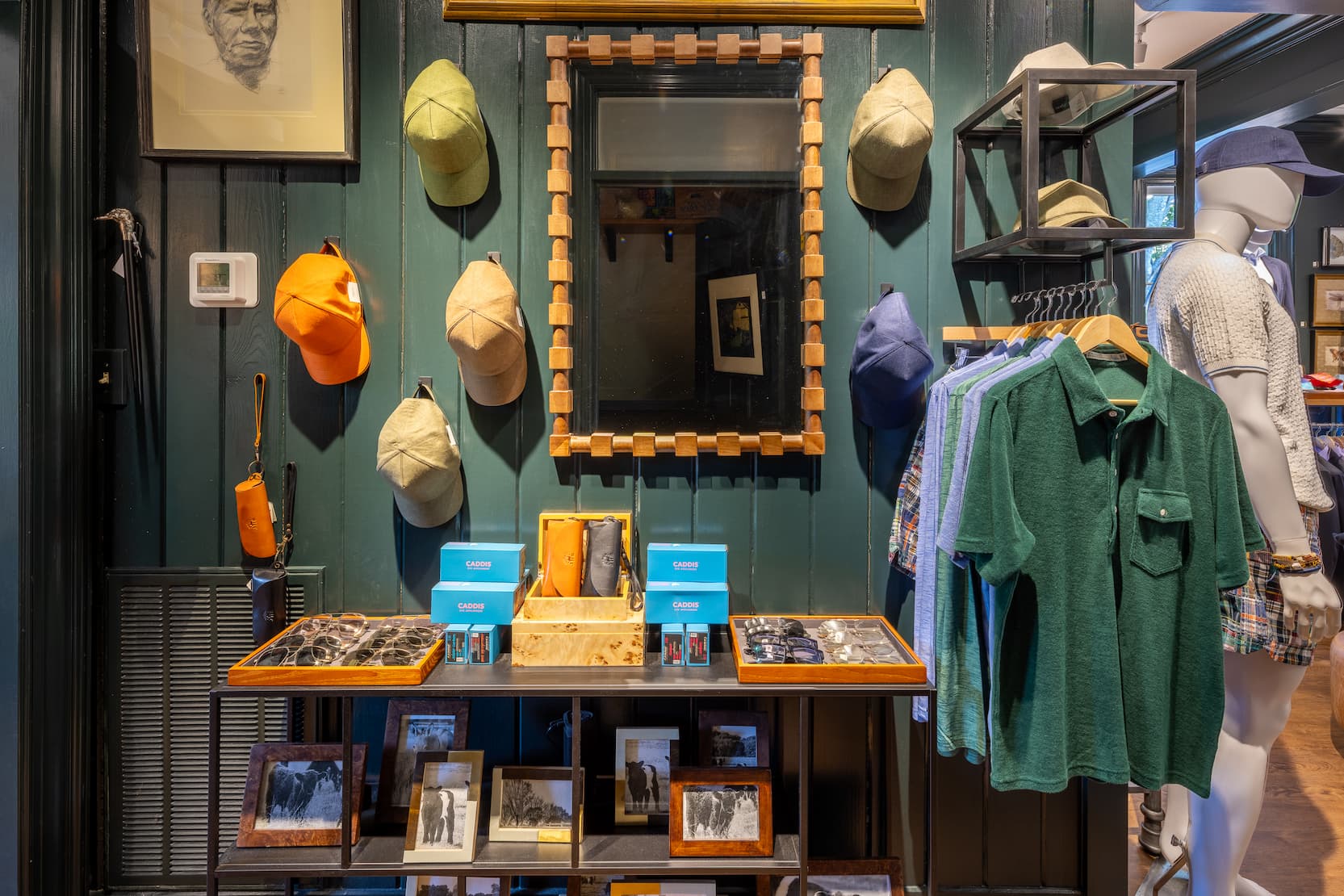 Upscale menswear display inside a boutique featuring deep green paneled walls, a geometric wood-framed mirror, and an assortment of colorful baseball caps mounted around it. Below, a display table showcases eyeglasses, leather accessories, and framed photographs. To the right, a rack of shirts includes a dark green knit polo and pastel button-downs, while a mannequin dressed in a textured cream polo and navy cap stands partially in view. Warm lighting and rich wood accents create a refined, classic atmosphere.