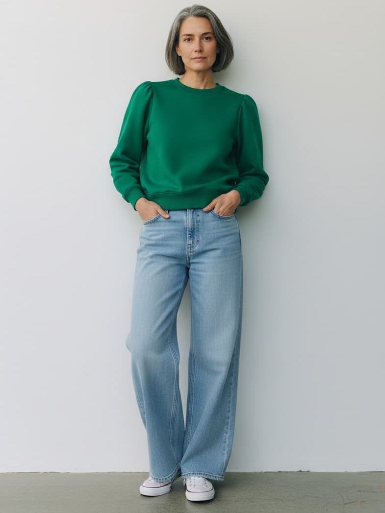 Woman in a green long-sleeve top and light blue wide-leg jeans, standing against a plain wall with hands in pockets and a relaxed, neutral expression.