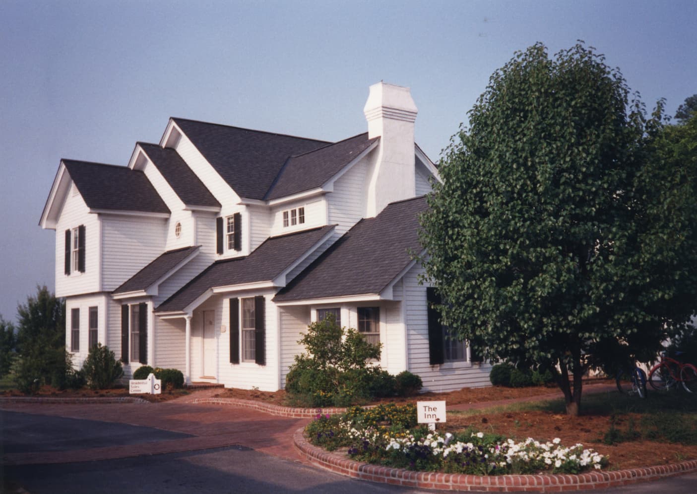 White clapboard inn with black shutters and a tall chimney, framed by a large leafy tree and a brick-edged flower bed with a small sign reading “The Inn.”