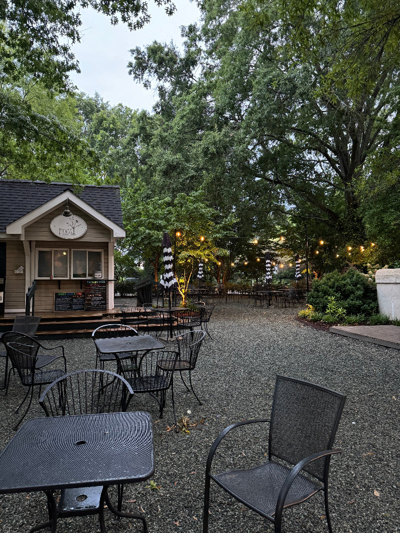 A quaint outdoor café with a small wooden building featuring a menu board and sign that reads "Food". The seating area has wrought-iron tables and chairs on a gravel ground. Black-and-white striped umbrellas are scattered around. Tall green trees surround the serene setting. Fearrington Village