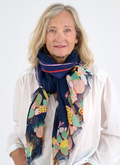 Woman with shoulder-length gray hair wears a white blouse and a navy scarf with colorful botanical patterns against a light background.