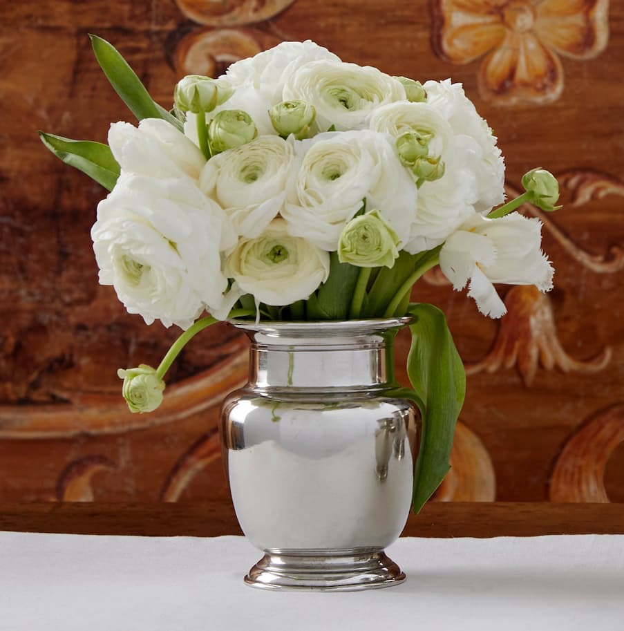 A polished pewter vase holds a compact arrangement of white ranunculus with green buds and a few long leaves, set against a warm, patterned wooden background.