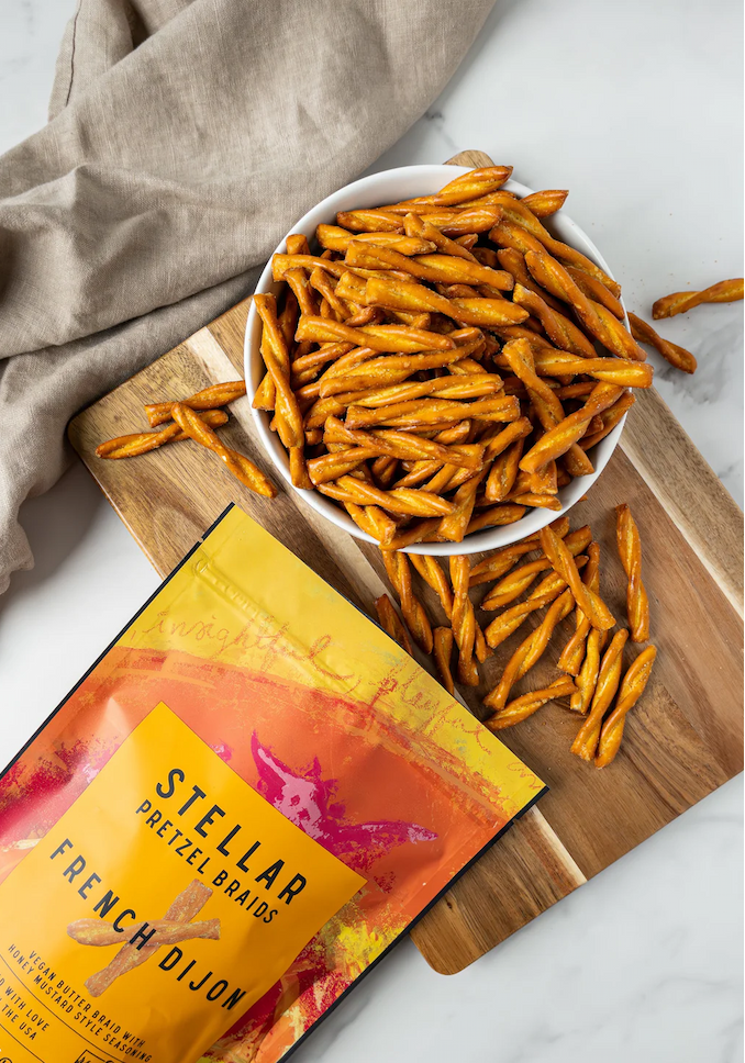 Bowl of Stellar Snacks French Dijon pretzel braids on a wooden board with scattered pretzels and the colorful snack bag beside it.
