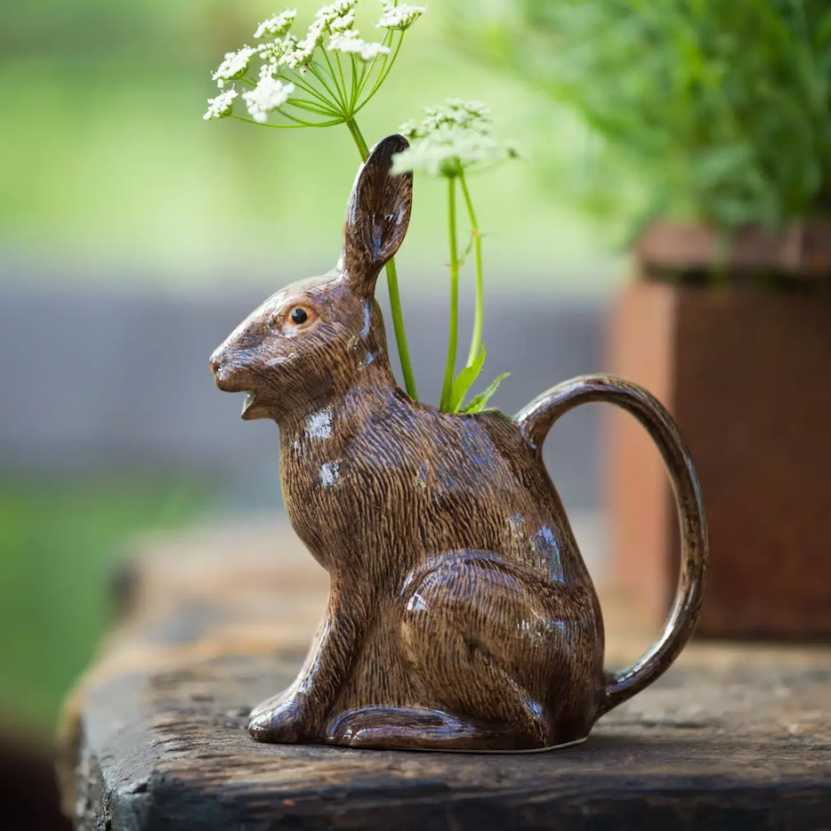 Brown ceramic rabbit-shaped vase sits on a rustic wooden surface holding delicate white wildflowers, with a soft green garden background.