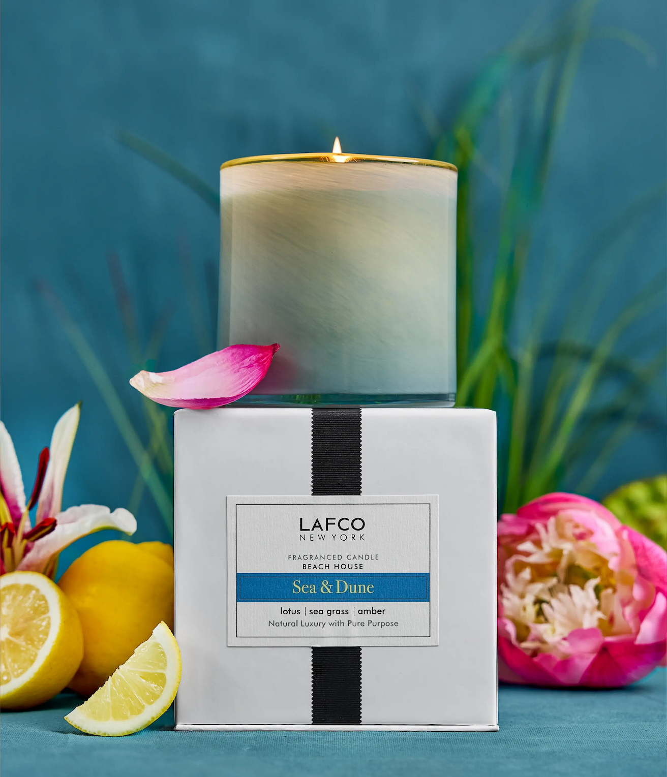 Lit LAFCO candle in a frosted glass vessel set atop its box, surrounded by citrus slices and flowers against a soft blue background.