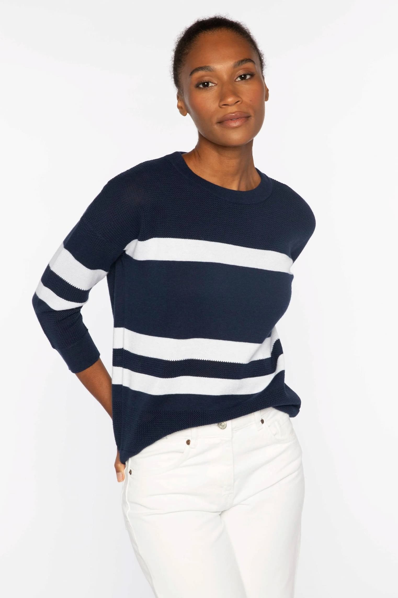 Woman wearing a navy knit sweater with bold white horizontal stripes, paired with light trousers against a neutral background.