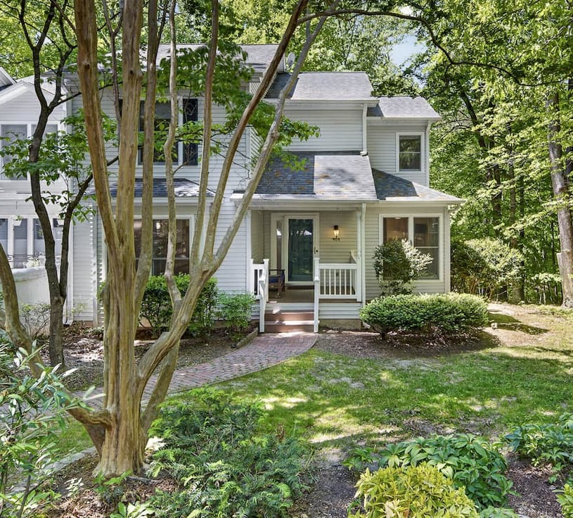 A charming two-story home with light siding sits nestled among trees, featuring a small front porch, brick walkway, and landscaped yard in a peaceful, wooded setting.