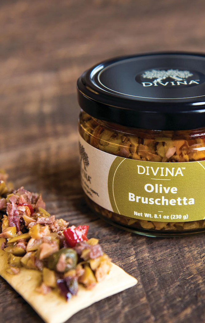A jar of Divina Olive Bruschetta sits on a wooden surface next to a cracker topped with the olive bruschetta mixture. Fearrington Village