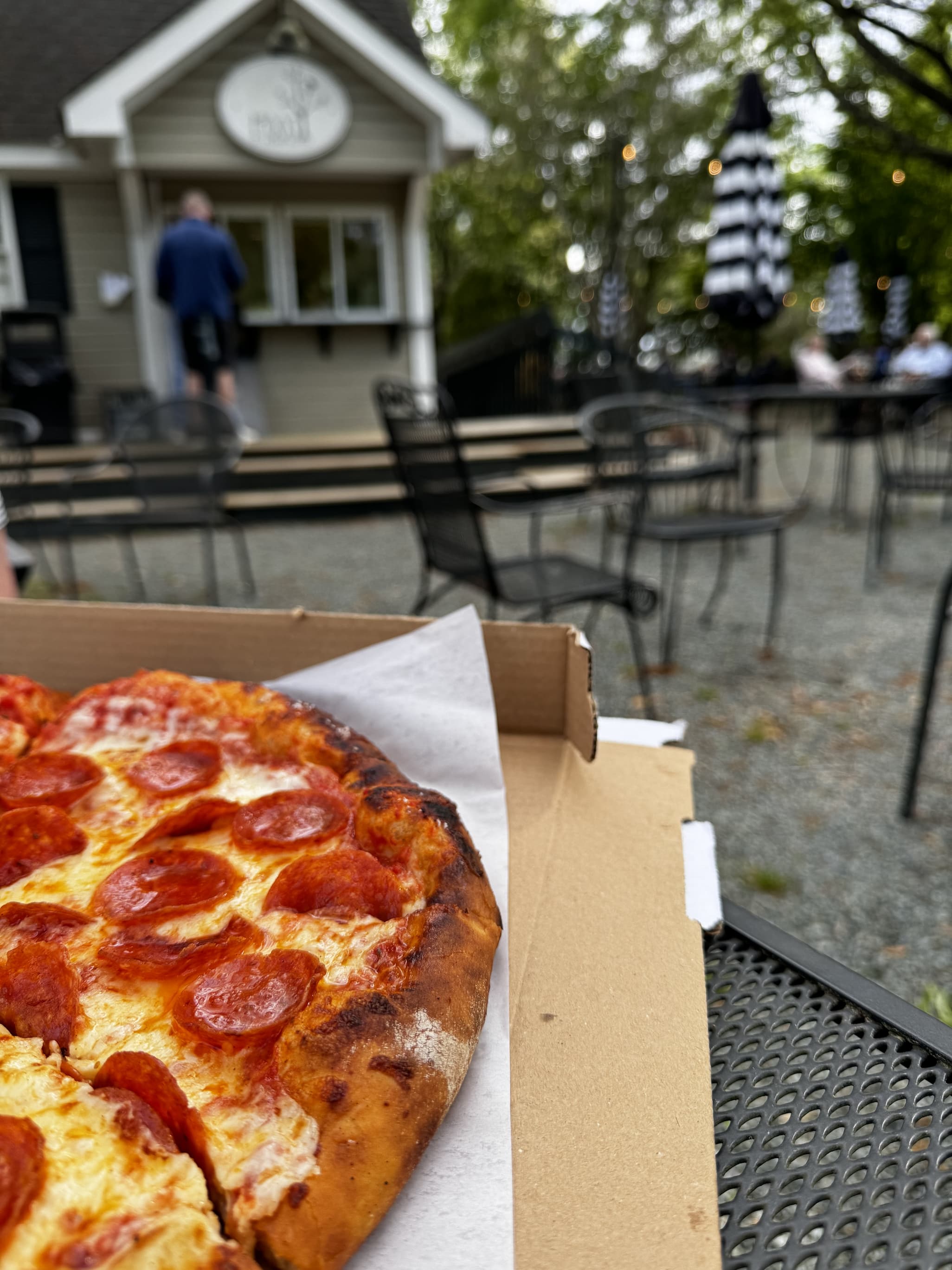 Close-up of a pepperoni pizza in an open box on a metal patio table, with a blurred outdoor café setting in the background. Behind it, a small food stand and scattered tables with black-and-white striped umbrellas sit among trees, suggesting a casual, relaxed dining area.