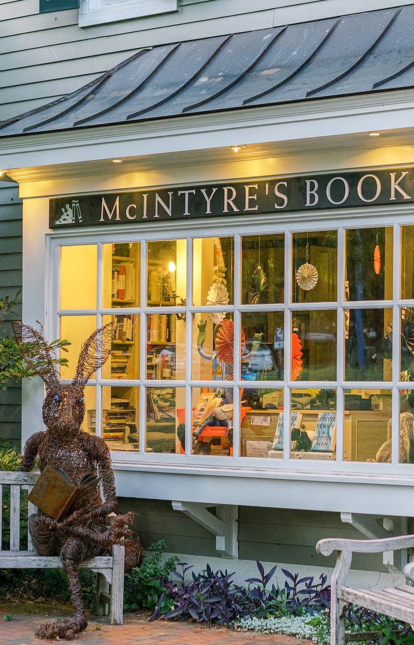 A cozy bookstore storefront with a sign reading “McIntyre’s Books.” Warm light glows through the windows filled with books and displays, while a whimsical sculpture of a rabbit reading a book sits on a bench outside.