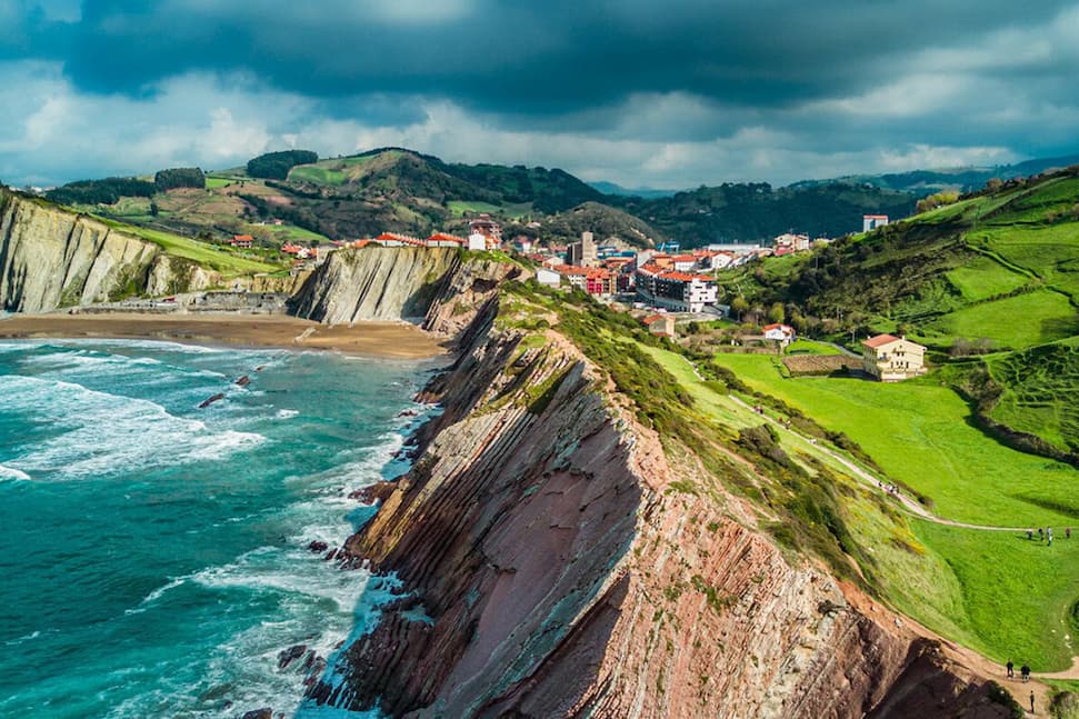 Dramatic coastal cliffs rise above turquoise waves along Spain’s rugged northern shoreline. Bright green hills, winding footpaths, and scattered farmhouses lead toward a seaside village nestled between the cliffs and rolling countryside, beneath dark, stormy clouds.