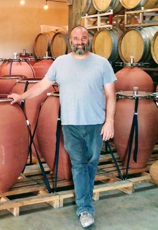 Winemaker standing in a cellar beside large terracotta amphorae, with stacked wooden wine barrels in the background, showcasing traditional winemaking craftsmanship.