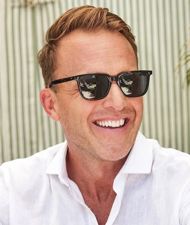 Man wearing black sunglasses and a white linen shirt, smiling outdoors in bright natural light against a softly striped background. Fearrington Village.