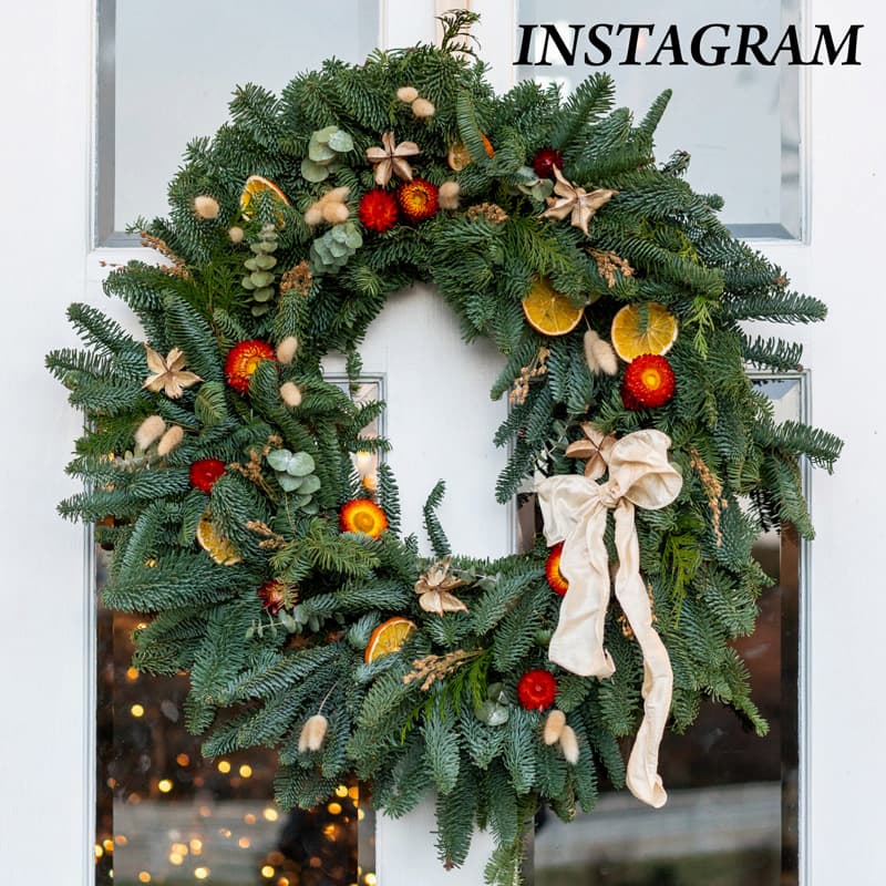 A festive wreath made of green pine branches, dried orange slices, red and yellow accents, star anise, and a cream-colored ribbon hangs on a white door. The word "INSTAGRAM" appears in the top right corner. Fearrington Village