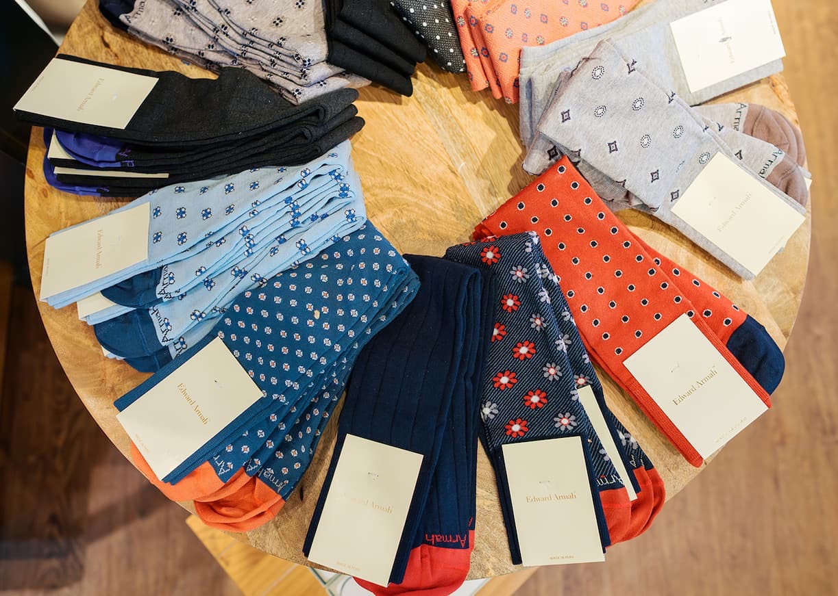 Round wooden display table topped with neatly folded men’s dress socks in assorted colors and patterns, each pair stacked with branded tags, photographed from above inside a boutique.