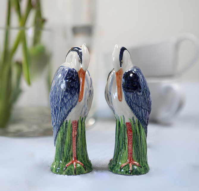 Pair of hand-painted ceramic bird figurines facing each other, with blue wings, orange beaks, and red legs, set on a table beside a teacup.