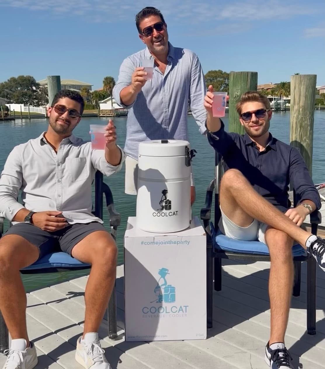 Three men seated by the water raising drinks around a CoolCat beverage cooler, enjoying a sunny, relaxed outdoor setting.
