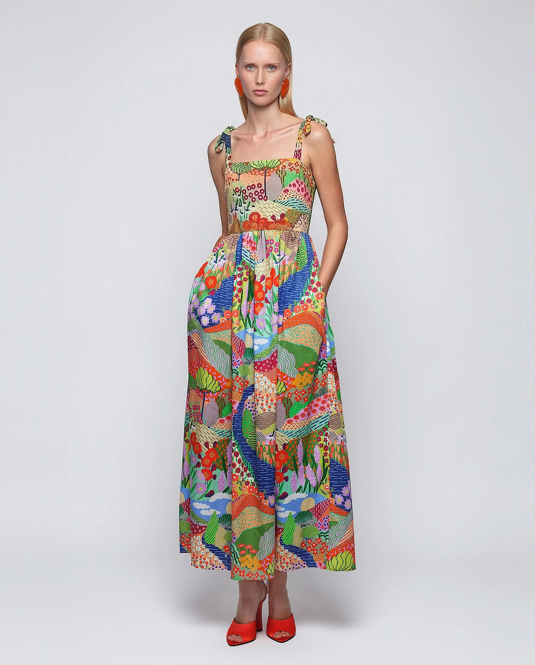 A model stands against a neutral backdrop wearing a colorful, patterned midi dress with tie straps, paired with bold red heels and statement earrings.