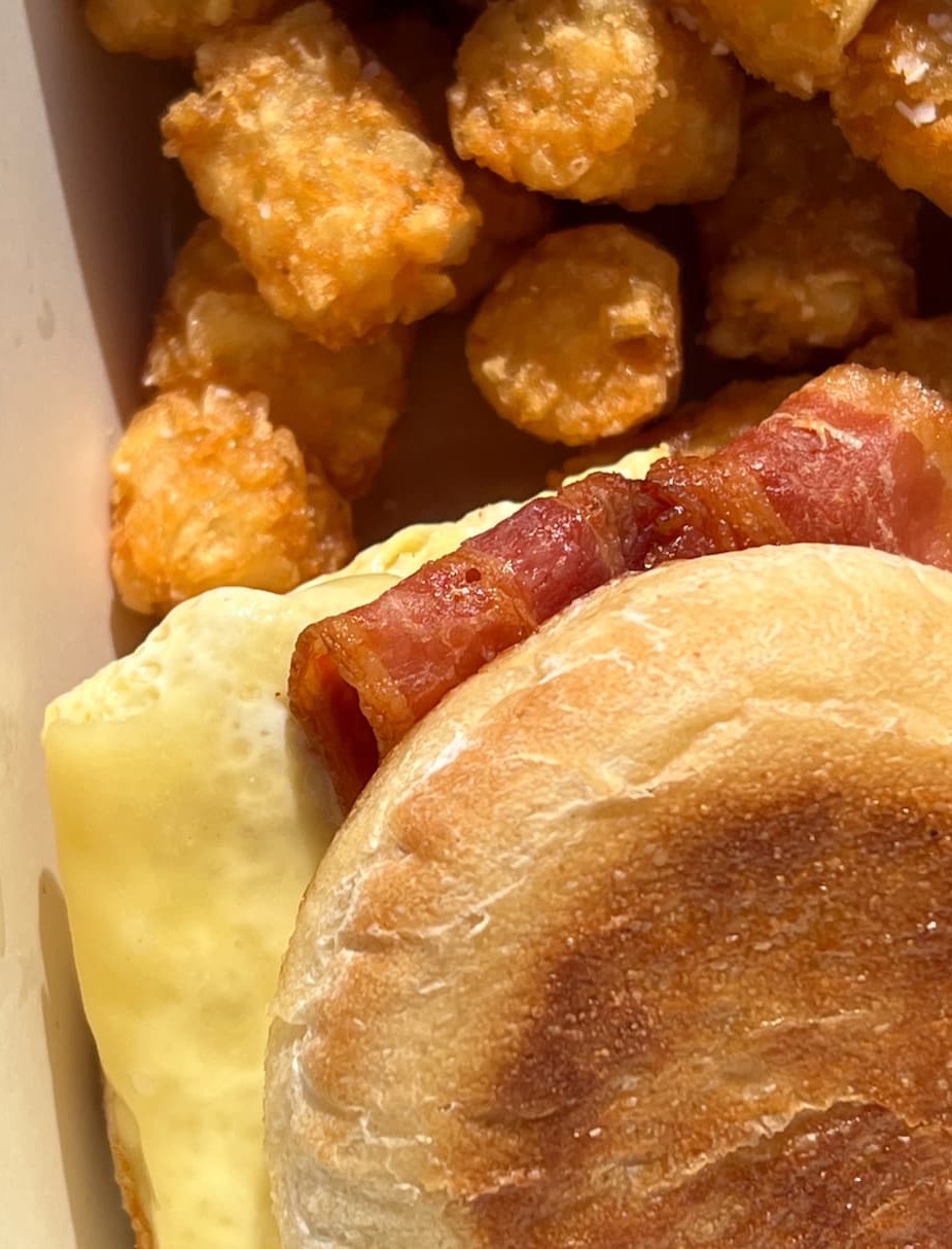 Close-up of a breakfast sandwich with a toasted bun, egg, and crispy bacon, served alongside a box of golden tater tots.