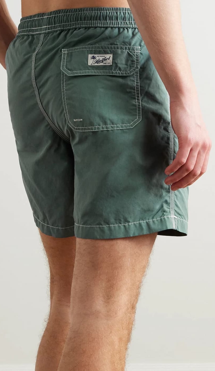 Close-up of green swim trunks with an elastic waistband and contrast stitching, featuring a back flap pocket with a branded label for a casual, classic look.