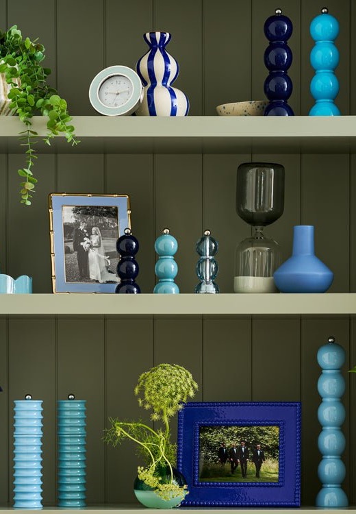 Built-in shelves styled with decorative objects in blue and green tones, including vases, candle holders, a clock, framed photos, and trailing greenery against a muted green backdrop.