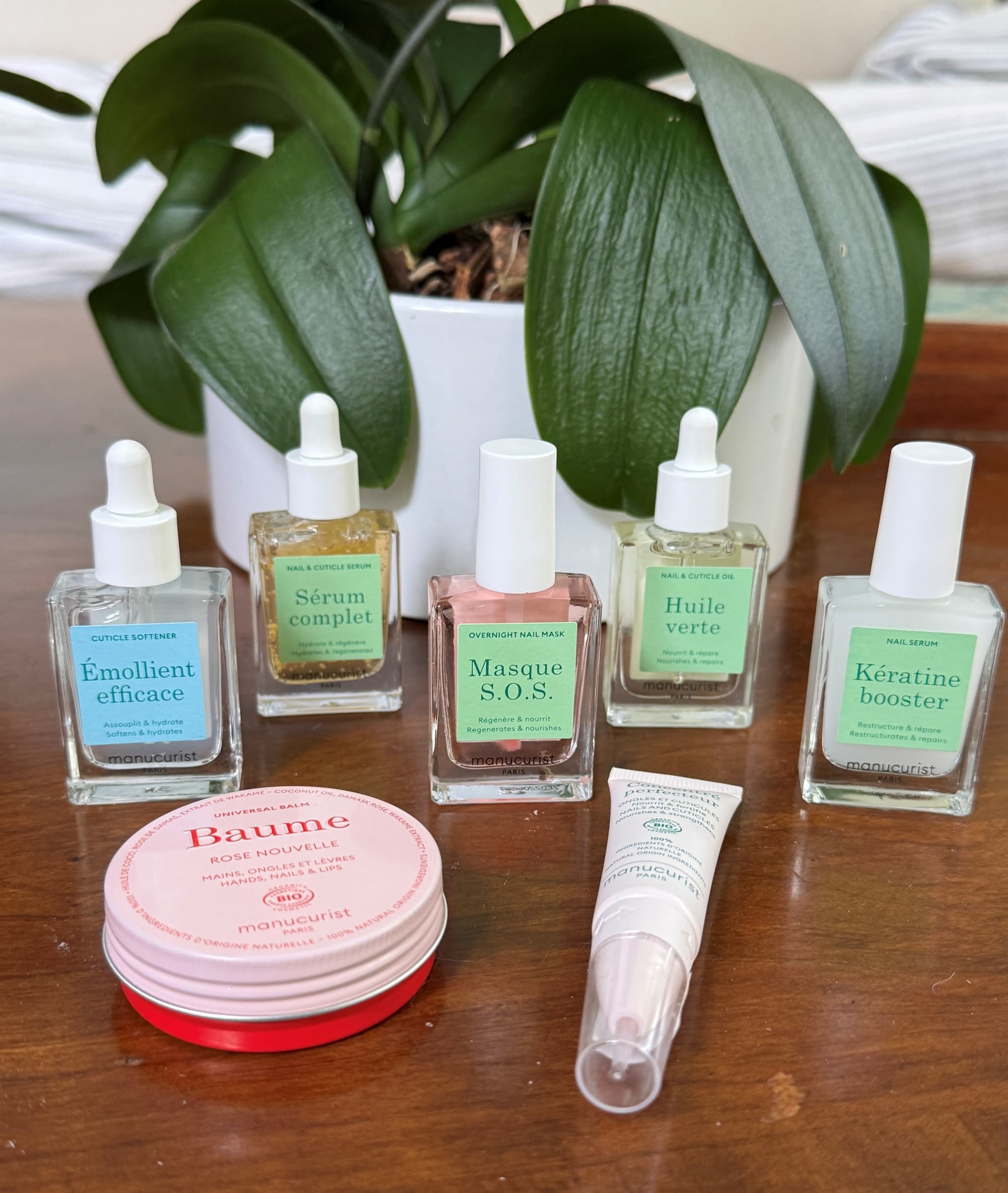 Assortment of Manucurist nail care products arranged on a wooden surface, including cuticle softener, serums, overnight mask, nail oil, keratin booster, and a rose balm, with a potted green orchid in the background.