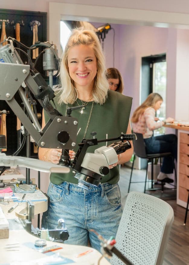 A smiling person stands at a jeweler’s workbench, holding specialized equipment, with tools and machinery in the foreground and others working at benches in a bright studio behind them.