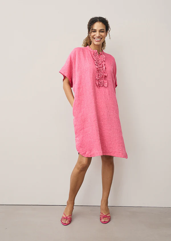 Smiling woman wearing a bright pink linen shift dress with ruffled detailing along the neckline and matching pink sandals, standing against a clean neutral backdrop.