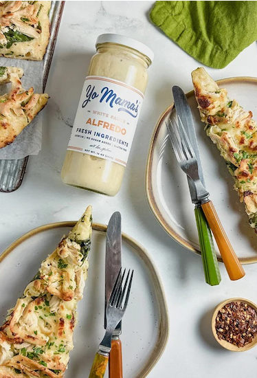 Flat-lay of a jar of Yo Mama’s Alfredo sauce surrounded by plated slices of cheesy flatbread, with forks and knives on neutral-toned dishes, set on a light surface with a green cloth and small bowl of seasoning nearby.