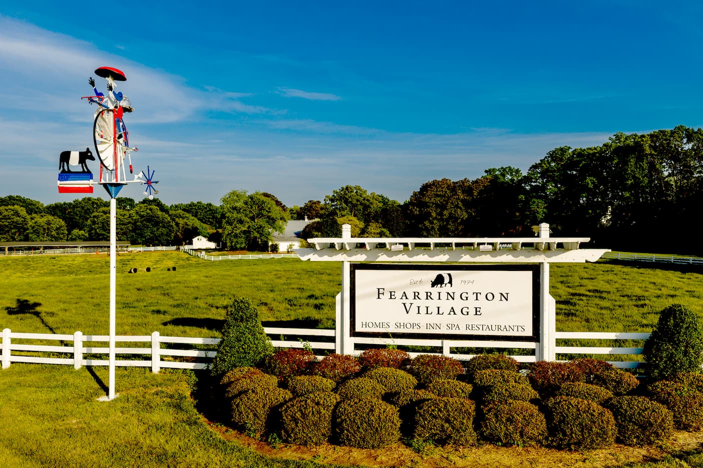Entrance sign for Fearrington Village set against a green pasture with white fencing, landscaped shrubs, and a decorative wind sculpture featuring a black cow; clear blue sky overhead.