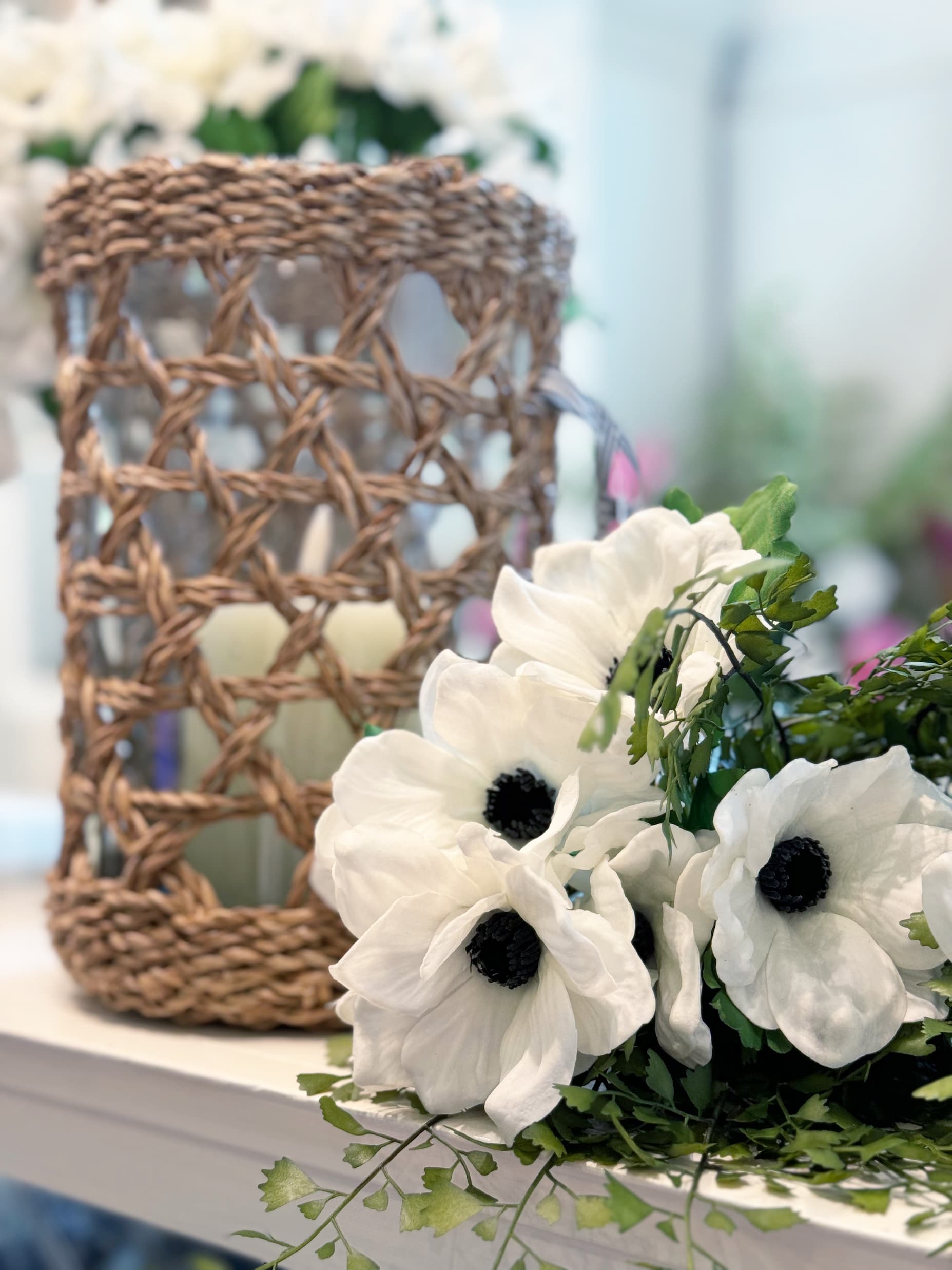 White anemone flowers with dark centers arranged beside a woven rattan hurricane candle holder on a soft white tabletop. Delicate greenery and blurred floral accents in the background create a calm, natural spring-inspired display.