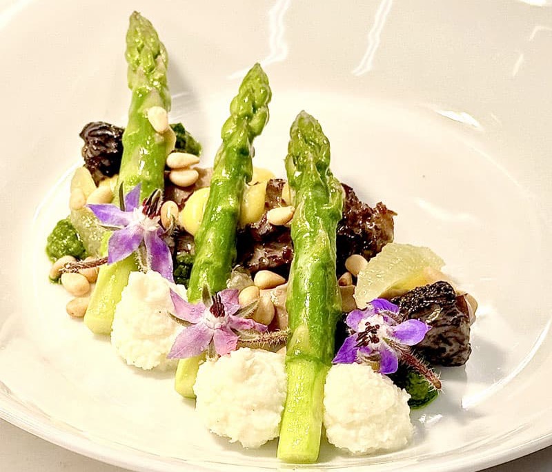 Plated dish featuring three upright asparagus spears over dollops of creamy purée, topped with sautéed mushrooms, pine nuts, and small yellow vegetable pieces. Garnished with purple edible flowers and a green herb sauce on a white plate.