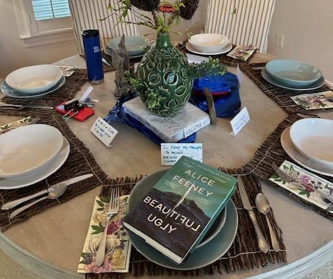 Dining table set for a gathering with plates and silverware, a green ceramic vase centerpiece, and a copy of Beautiful Ugly by Alice Feeney placed at one setting.