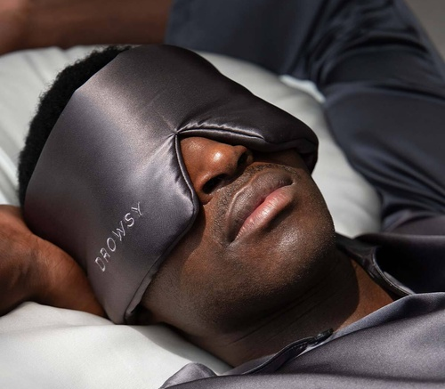 Person lying on a pillow wearing a dark satin sleep mask that contours around the nose, resting with eyes covered and relaxed expression.