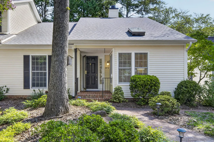 Single-story cottage with white siding, black shutters, a brick entryway, and mature landscaping beneath tall shade trees. Fearrington Village.