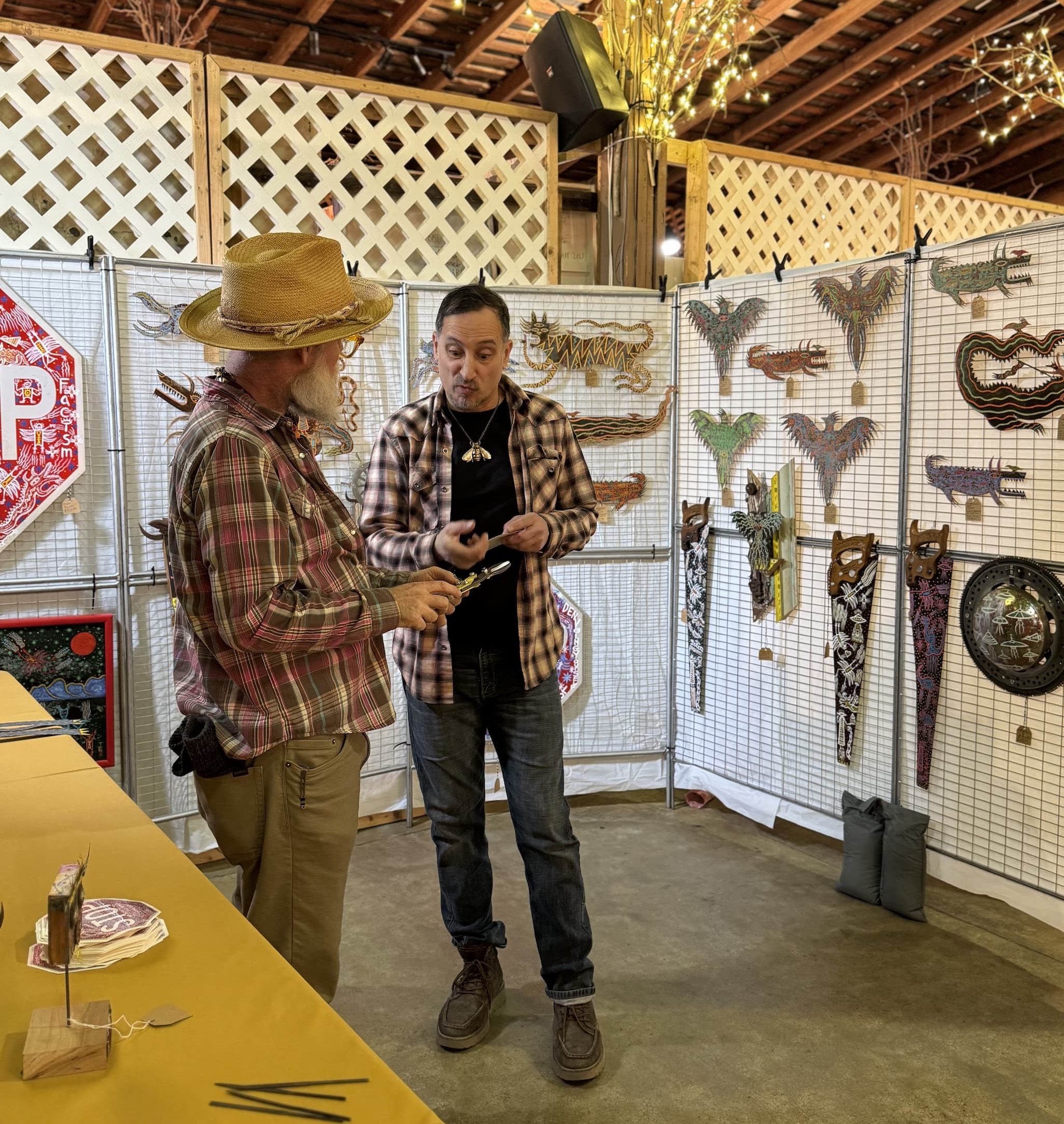 Two men talk inside a rustic barn at an art fair, standing beside a table and a display wall of colorful folk art sculptures and wall pieces. One man wears a straw hat and plaid shirt, while the other gestures as he explains an item, with lattice walls and string lights overhead.
