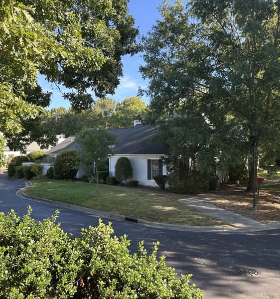 A white house with dark roof sits on a tree-lined corner in a suburban neighborhood. The street curves around the house, and sunlight filters through large green trees, casting shadows on the pavement and yard. Fearrington Village
