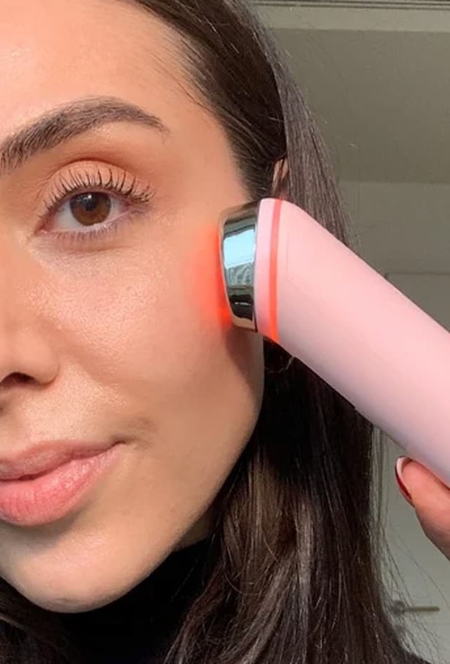 Woman using a handheld facial device emitting red light against her cheek during a skincare treatment.
