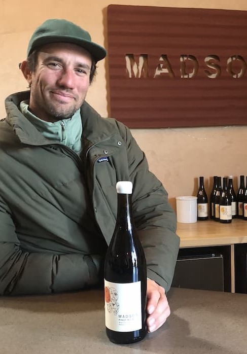 Man in a green cap and jacket stands behind a tasting counter, holding a bottle of wine. Behind him, a wall sign partially reads “MADSO,” with several wine bottles displayed on a shelf in a warm, casual winery setting.