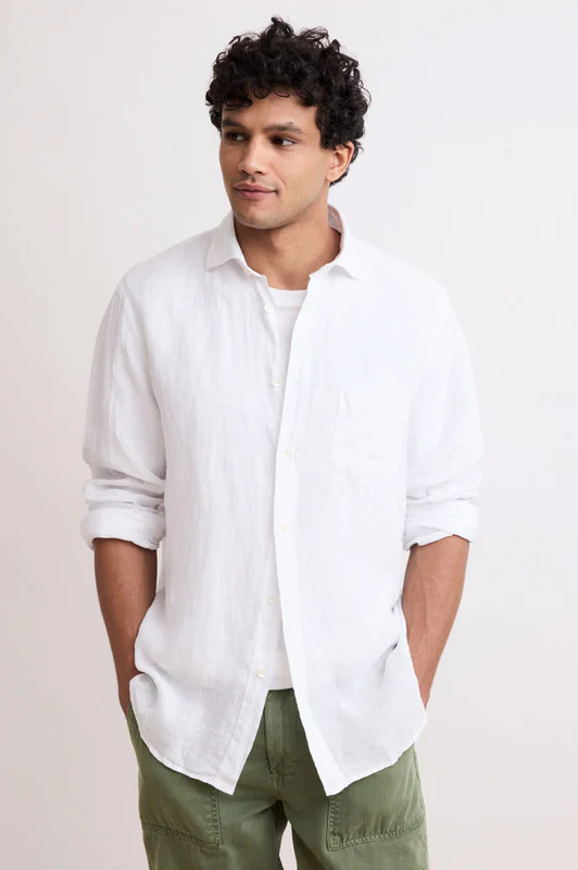 Man wearing a relaxed white button-down shirt and olive green pants, standing against a neutral background with hands in pockets.