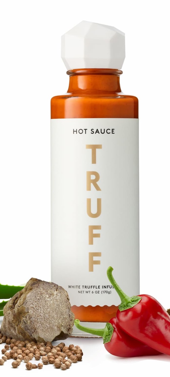 Bottle of TRUFF White Truffle Infused Hot Sauce with a faceted white cap, styled with fresh red chili peppers, a truffle, coriander seeds, and aloe leaves on a white background.