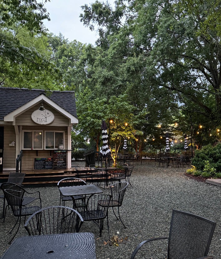 A quaint outdoor café with a small wooden building featuring a menu board and sign that reads "Food". The seating area has wrought-iron tables and chairs on a gravel ground. Black-and-white striped umbrellas are scattered around. Tall green trees surround the serene setting. Fearrington Village