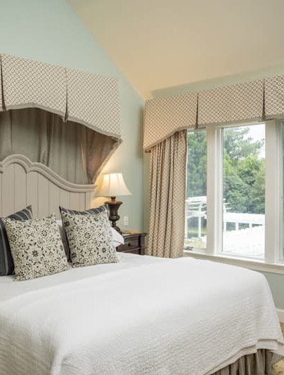Bright guest bedroom with a neatly made white bed, patterned accent pillows, a curved headboard, soft green walls, and a large window dressed with tailored valances overlooking a white fence and trees.