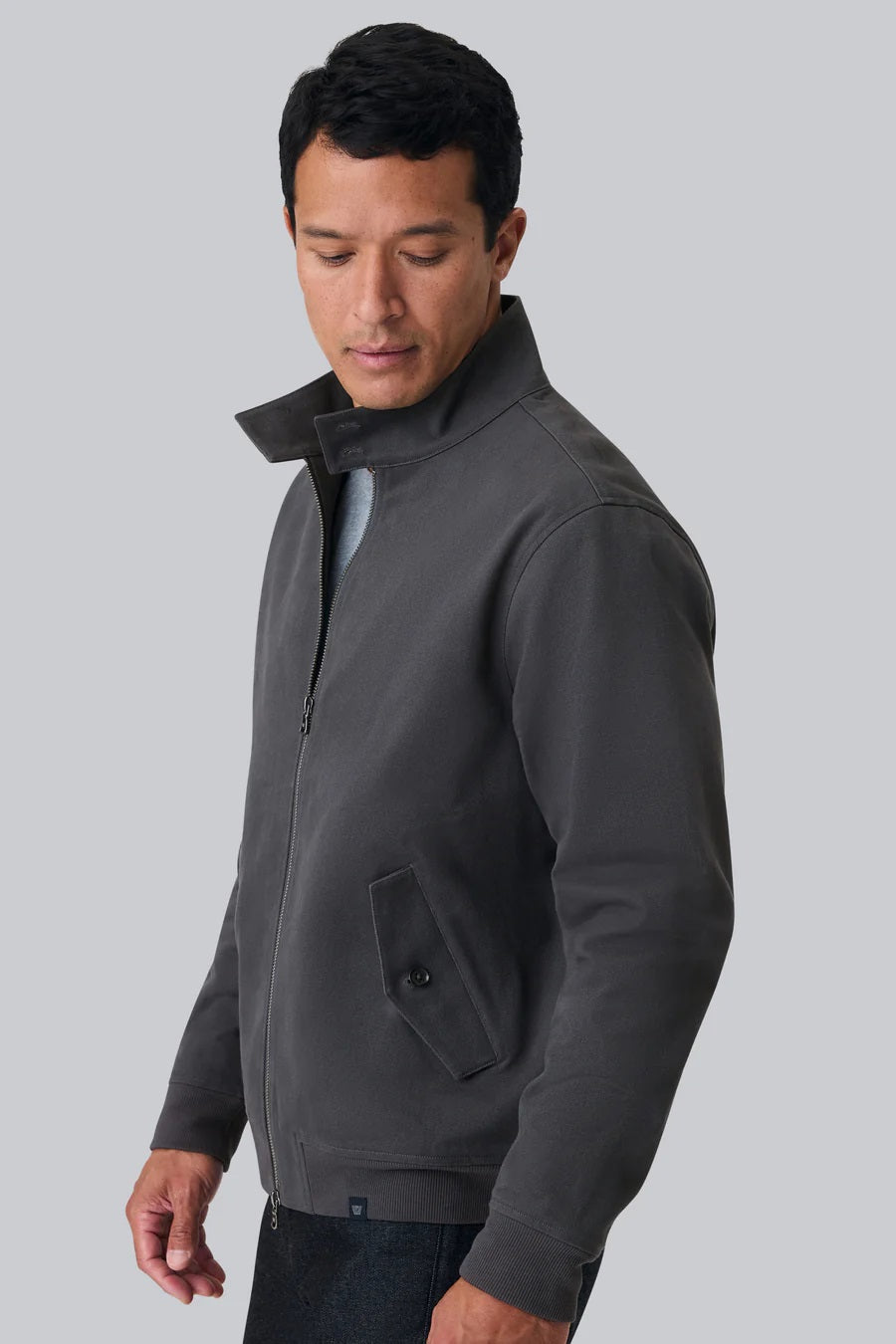 Man wearing a charcoal zip-front jacket with stand collar looks down against a light gray background.