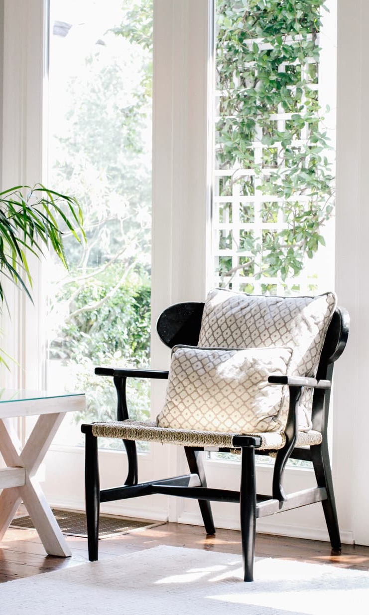 Black wood accent chair with woven seat and patterned pillows beside tall sunlit windows overlooking greenery and climbing vines.