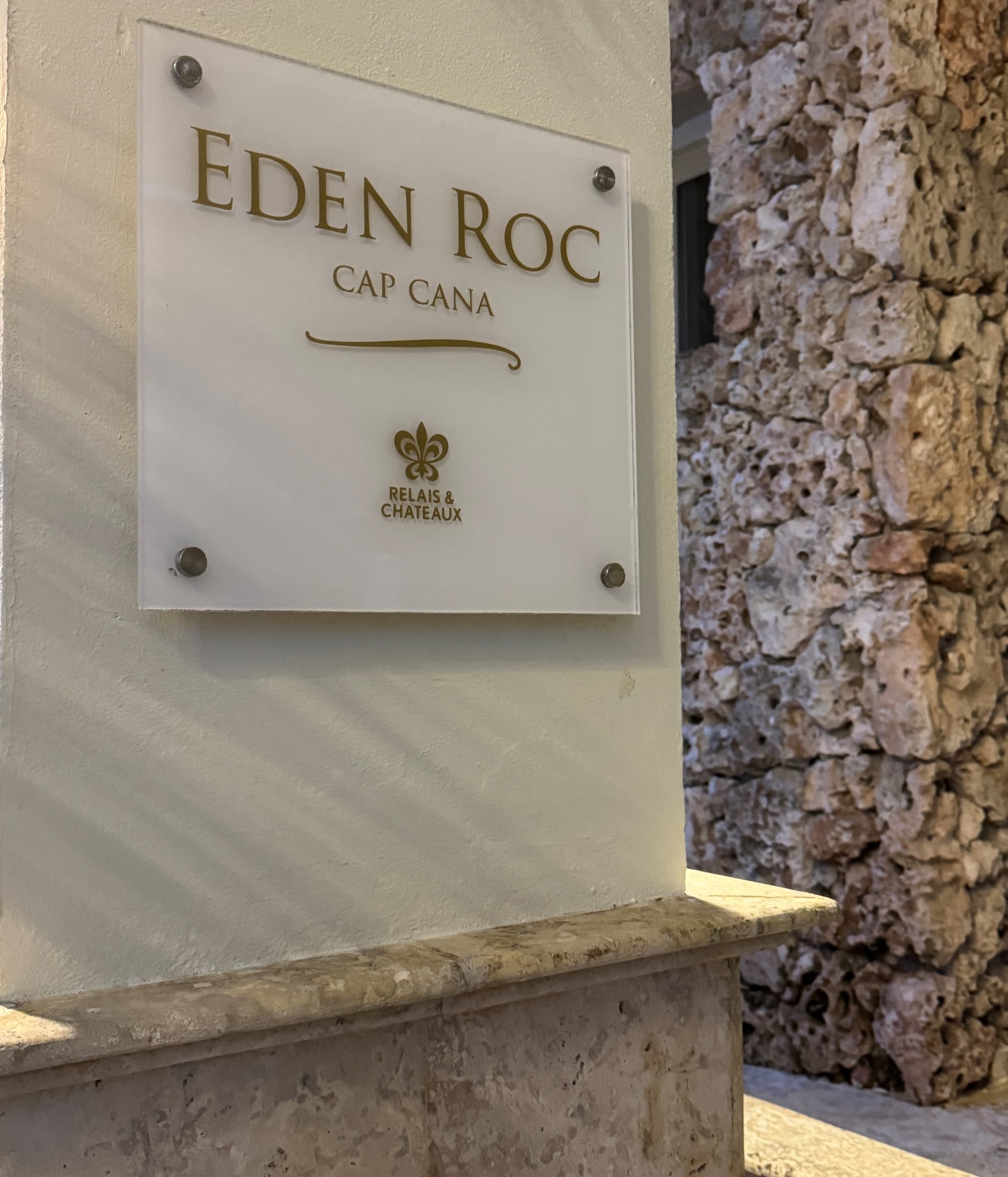 Close-up of a clear acrylic wall plaque reading “Eden Roc Cap Cana” with the Relais & Châteaux emblem, mounted on a white stucco wall with metal standoffs; textured stone wall visible to the right and a ledge below, lit by warm natural light.